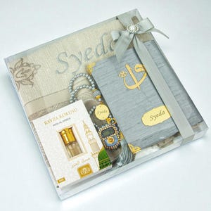 Op de afbeelding: Een geschenkset in een doorzichtige doos, inclusief een gebedskleed, een boek, een digitale gebedskralenteller en een klein flesje parfum. De set is vastgebonden met een grijs lint en strik. De items hebben de naam "Syeda" erop.