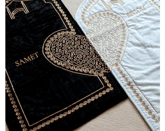 May include: Two prayer rugs, one black and one white, with gold embroidered details. The black rug has the name "SAMET" embroidered in gold. Both rugs feature a heart-shaped design with intricate gold patterns and a decorative border.