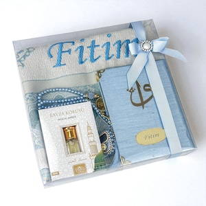 Op de afbeelding: Een geschenkset in een transparante doos, met een lichtblauw gebedskleed geborduurd met "Fitim", een blauwe Koran, blauwe gebedskralen en een klein parfumflesje. De doos is vastgebonden met een lichtblauw lint en een decoratieve sluiting.