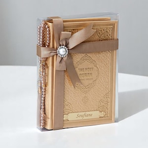 May include: A gold-colored Holy Quran book with the text "THE HOLY QURAN" and "BASIC-ENGLISH" is displayed in a clear box. A matching rosary and a brown ribbon with a pearl embellishment are included. The name "Soufiane" is printed on the book.