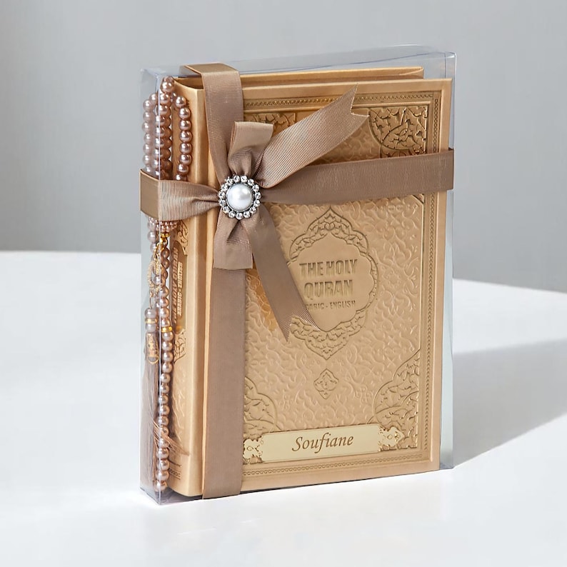 May include: A gold-colored Holy Quran book with the text "THE HOLY QURAN" and "BASIC-ENGLISH" is displayed in a clear box. A matching rosary and a brown ribbon with a pearl embellishment are included. The name "Soufiane" is printed on the book.