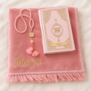 Op de afbeelding: Een roze fluwelen gebedskleed met de naam Marya in goud geborduurd, vergezeld door een bijpassende roze kralen gebedsketting met een gouden bedel gegraveerd met Marya, en een roze Heilige Koran met gouden letters en de naam Marya.