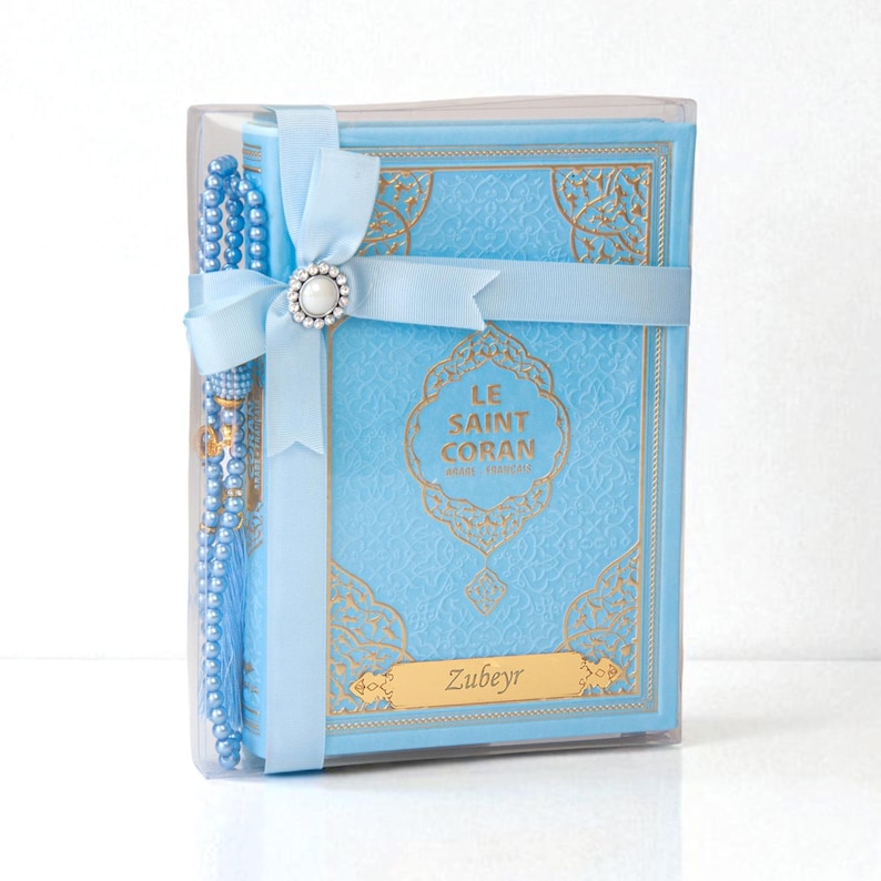May include: A light blue book with gold lettering and a matching rosary, presented in a clear box. The book is titled "LE SAINT CORAN" and includes the name "Zubeyr" on a gold plate. A light blue ribbon with a pearl-centered embellishment secures the set.