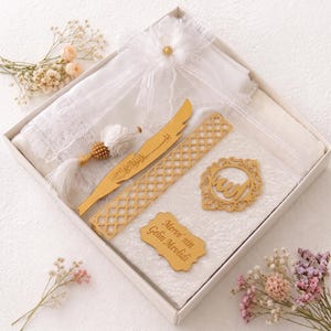 May include: A white gift box containing a white lace-trimmed fabric, a white prayer bead set with a gold tassel, a gold feather-shaped bookmark, a gold lattice-patterned bookmark, a gold circular calligraphy ornament, and a gold plaque with text.