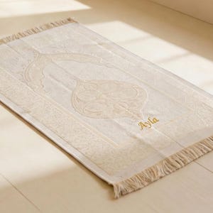 May include: Cream-colored prayer rug with an ornate central design and a fringed edge. The rug features the name "Ayla" embroidered in gold thread. The rug is rectangular and lies on a light-colored wooden surface.