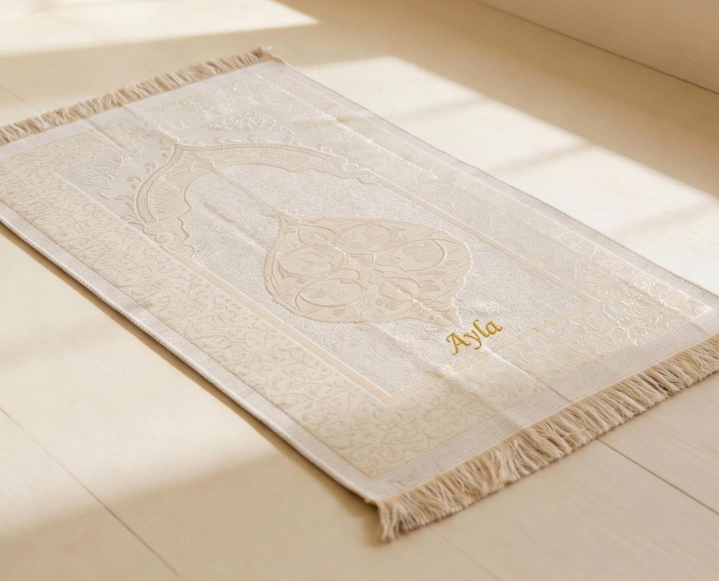 May include: Cream-colored prayer rug with an ornate central design and a fringed edge. The rug features the name "Ayla" embroidered in gold thread. The rug is rectangular and lies on a light-colored wooden surface.