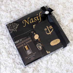 May include: A black gift box with a black ribbon and a decorative jewel. Inside are a black prayer mat with gold embroidery, a black book with gold lettering, a small bottle, and a beaded bracelet. The name "Nasif" is written in gold.