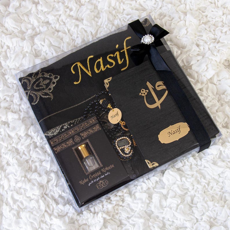 May include: A black gift box with a black ribbon and a decorative jewel. Inside are a black prayer mat with gold embroidery, a black book with gold lettering, a small bottle, and a beaded bracelet. The name "Nasif" is written in gold.