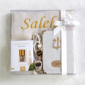 May include: A gift set in a clear box, featuring a beige prayer rug embroidered with "Saleh" in gold, a pearl necklace, a small perfume bottle, a digital counter, and a white book with gold accents. The set is tied with a white ribbon.