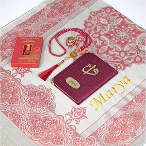 May include: A prayer mat with a pink and white floral design, a burgundy book with gold lettering, a red beaded rosary, and a small red box. The book is inscribed with the name "Marya" in gold. The items are arranged on a white surface.