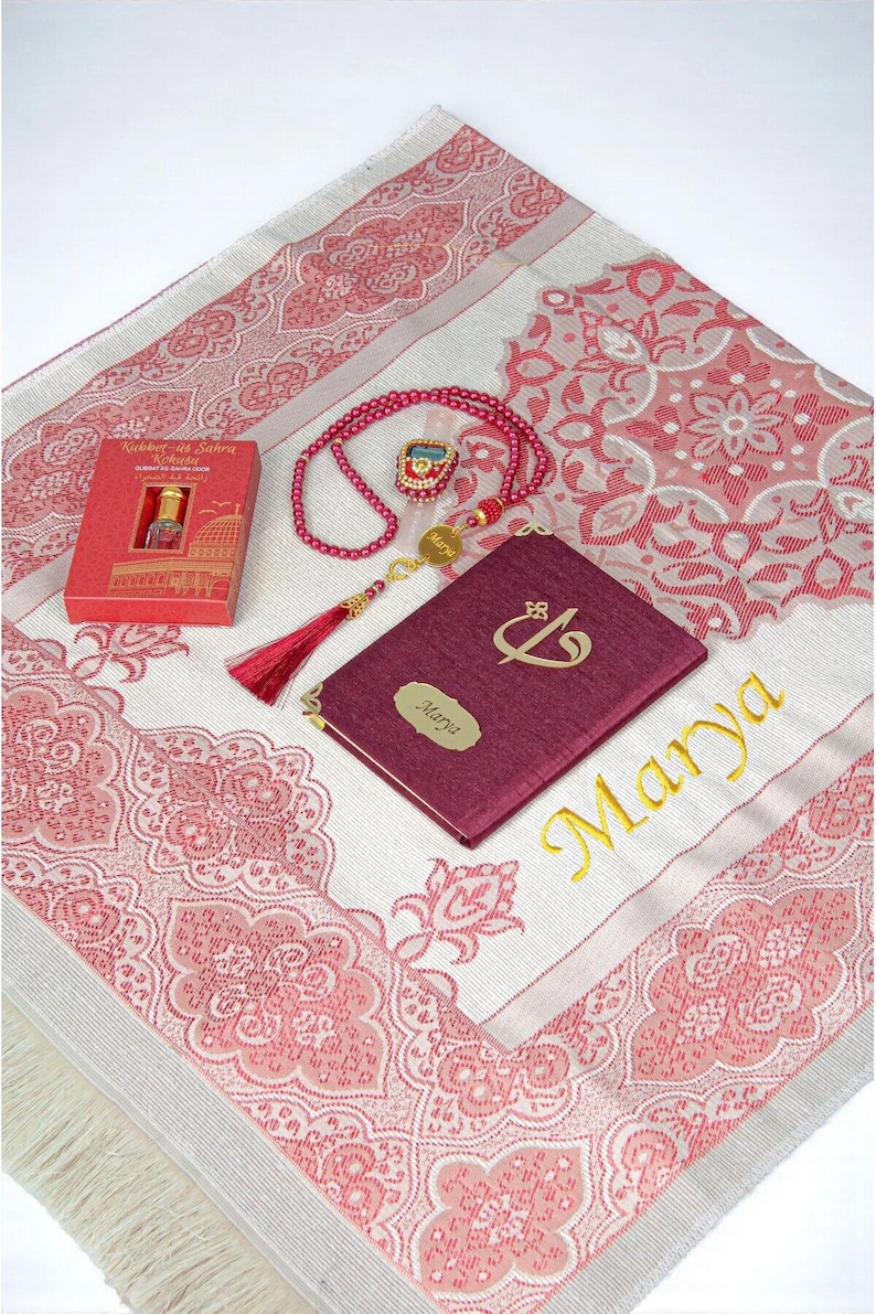 May include: A prayer mat with a pink and white floral design, a burgundy book with gold lettering, a red beaded rosary, and a small red box. The book is inscribed with the name "Marya" in gold. The items are arranged on a white surface.