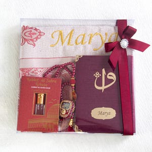 May include: A gift set in a clear box, featuring a prayer mat embroidered with "Marya" in gold, a burgundy book, a rosary, and a small perfume bottle. A burgundy ribbon with a pearl embellishment secures the set.