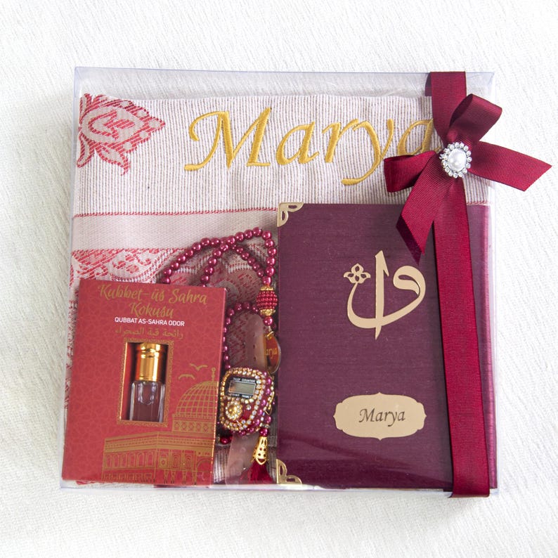 May include: A gift set in a clear box, featuring a prayer mat embroidered with "Marya" in gold, a burgundy book, a rosary, and a small perfume bottle. A burgundy ribbon with a pearl embellishment secures the set.