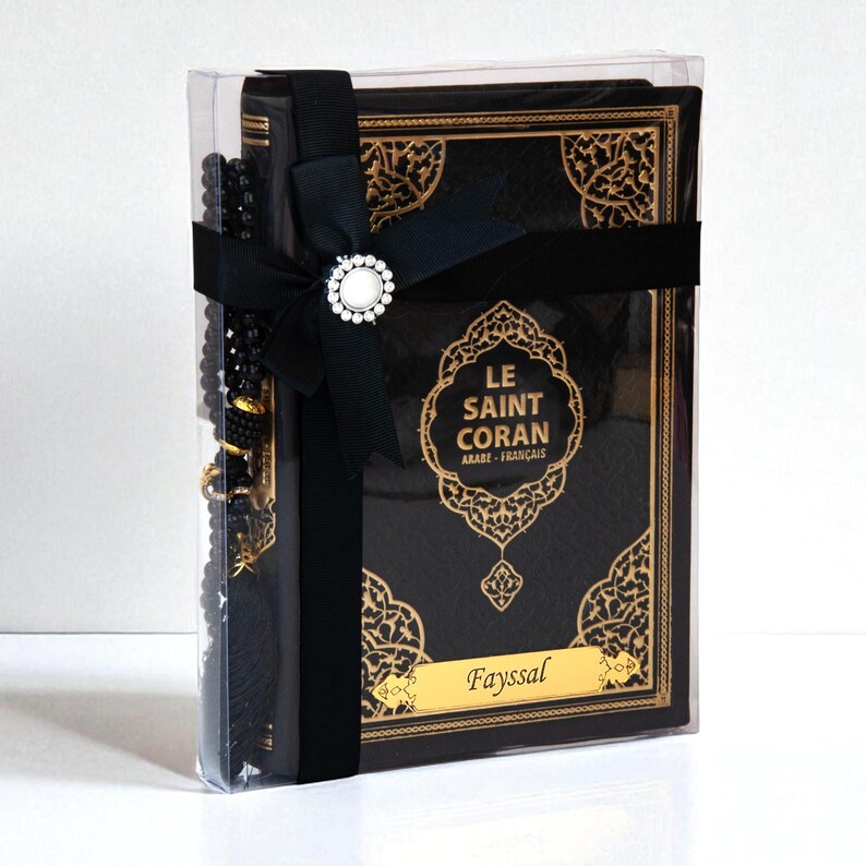 May include: A black book with gold accents and the text "LE SAINT CORAN ARABE-FRAN&Ccedil;AIS" is presented in a clear box, tied with a black ribbon and a decorative clasp. A black beaded strand is also included.
