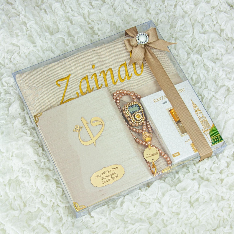 May include: A gift set in a clear box, featuring a beige prayer mat embroidered with "Zainab" in gold, a book with gold accents, a beaded rosary, and a small perfume box. The set is tied with a beige ribbon and a decorative clasp.