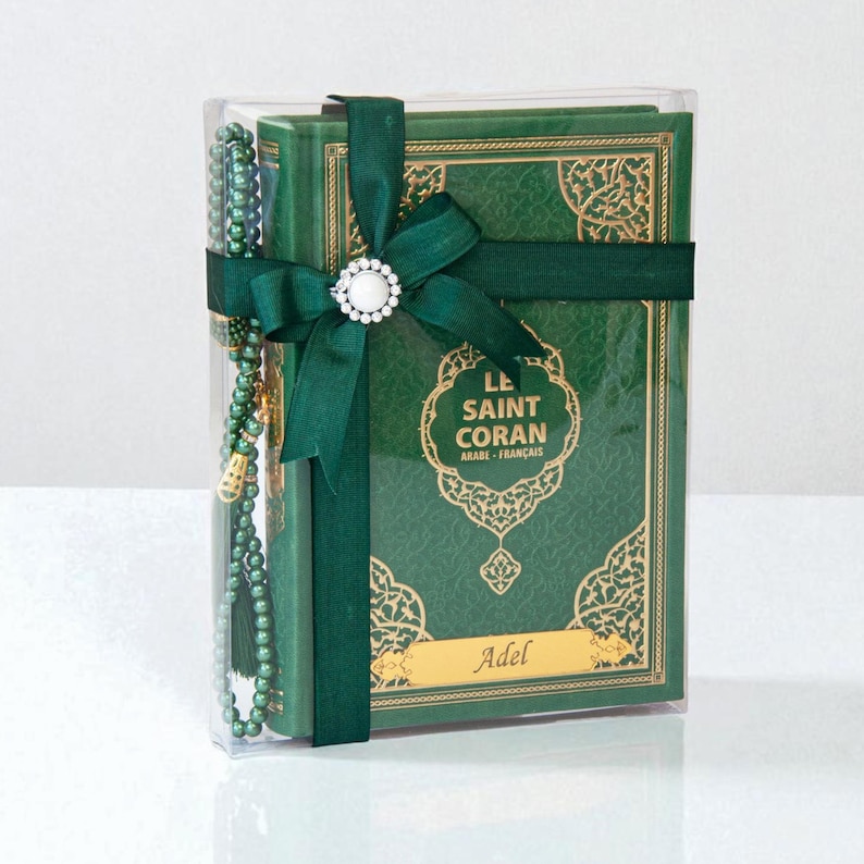 May include: A green book set, including a Quran, is presented in a clear box, tied with a dark green ribbon and a decorative white and silver embellishment. A matching green beaded prayer accessory is included. The book cover features gold filigree and the text "LE SAINT CORAN ARABE - FRAN&Ccedil;AIS".