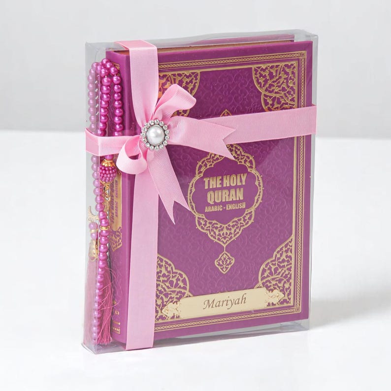 May include: A boxed set featuring a purple and gold embossed book titled "The Holy Quran" with Arabic and English text, a pink ribbon, and a beaded rosary. The set is wrapped in clear plastic.