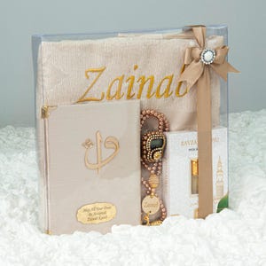 May include: A gift set in a clear box, featuring a beige prayer mat embroidered with "Zainab" in gold, a beige book with gold accents, a beaded digital counter, and a small perfume bottle. The box is tied with a beige ribbon and a decorative jewel.