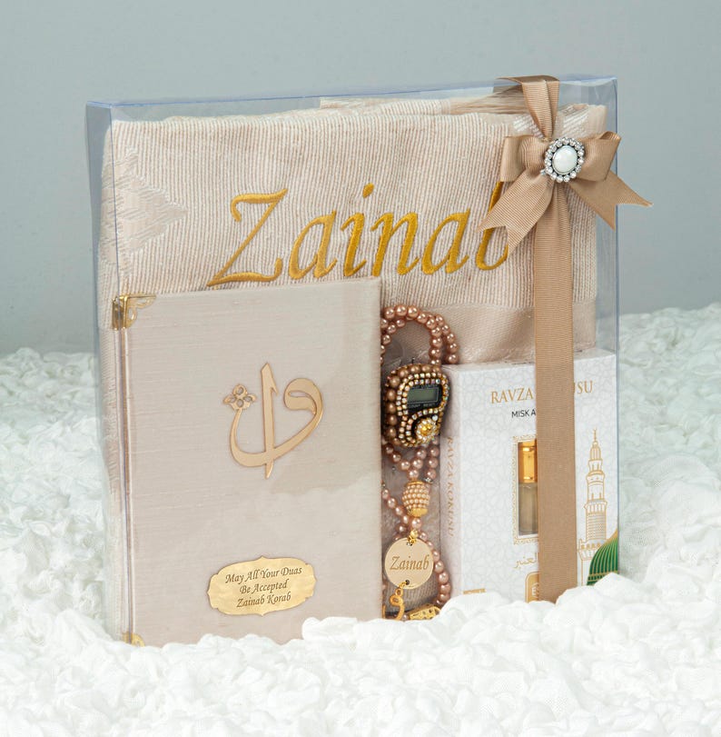 May include: A gift set in a clear box, featuring a beige prayer mat embroidered with "Zainab" in gold, a beige book with gold accents, a beaded digital counter, and a small perfume bottle. The box is tied with a beige ribbon and a decorative jewel.