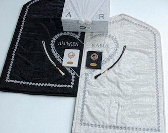 May include: A black and white prayer mat set with a decorative box. The black mat has the name "ALPEREN" embroidered in a heart shape. The white mat has the name "RABIA" embroidered in a heart shape. The box has the letters "A" and "R".