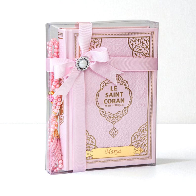 May include: A pink Quran set in a clear box, tied with a pink ribbon and a pearl-centered embellishment. The book has gold filigree and the text "LE SAINT CORAN ARABE - FRAN&Ccedil;AIS". A pink prayer bead set is included.