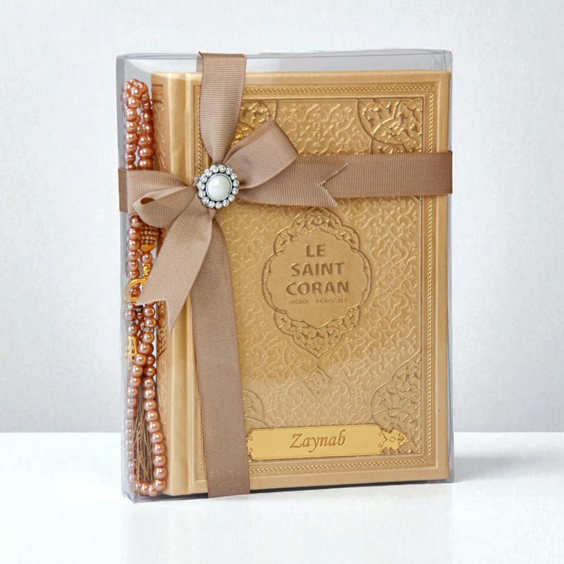 May include: A gold-colored book titled "LE SAINT CORAN" with a decorative embossed cover, presented in a clear box. A brown ribbon with a jeweled accent and a string of beads are wrapped around the book. The name "Zaynab" is printed on the book.