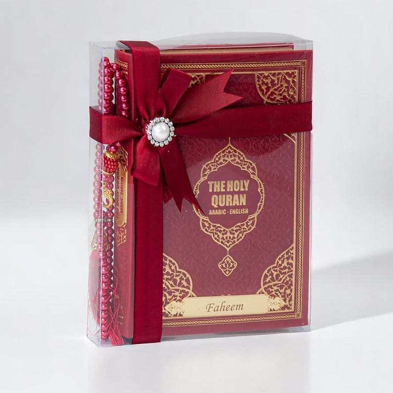 May include: A boxed set featuring a burgundy-colored Quran with gold filigree, a matching prayer bead strand, and a decorative bow with a pearl-centered embellishment. The text on the cover reads "THE HOLY QURAN ARABIC - ENGLISH" and includes the name "Faheem".