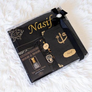 May include: A black gift box with a black scarf embroidered with "Nasif" in gold, a black prayer book with gold calligraphy, a black beaded rosary, and a small perfume bottle. The box is tied with a black ribbon and a decorative jewel.