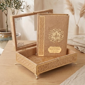 May include: A beige velvet box with a clear lid, open to reveal a matching book. The book has gold decorative accents and a gold plaque with text. The box is on a wooden surface.