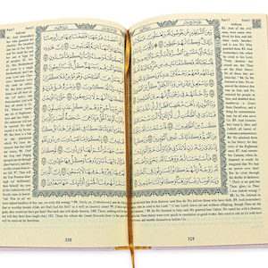 May include: Open book with Arabic script, featuring ornate borders and a gold ribbon bookmark. The pages display text with decorative elements. The book has a light pink cover and is open to pages 329 and 330.