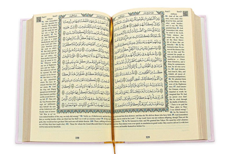 May include: Open book with Arabic script, featuring ornate borders and a gold ribbon bookmark. The pages display text with decorative elements. The book has a light pink cover and is open to pages 329 and 330.