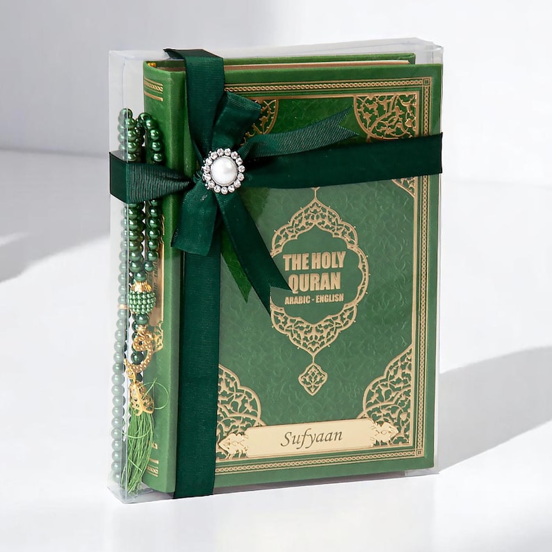 May include: A green book titled "THE HOLY QURAN ARABIC - ENGLISH" is wrapped with a dark green ribbon and a pearl-centered embellishment. A matching green beaded strand is included. The book is in a clear box. The name "Sufyaan" is printed on the book.
