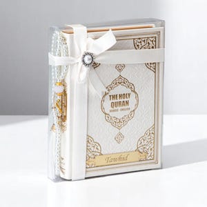 May include: A white and gold embossed book set, titled "The Holy Quran Arabic English," is presented in a clear box. A white ribbon with a pearl embellishment secures the set, which includes a prayer bead strand. The word "Tawhid" is inscribed.
