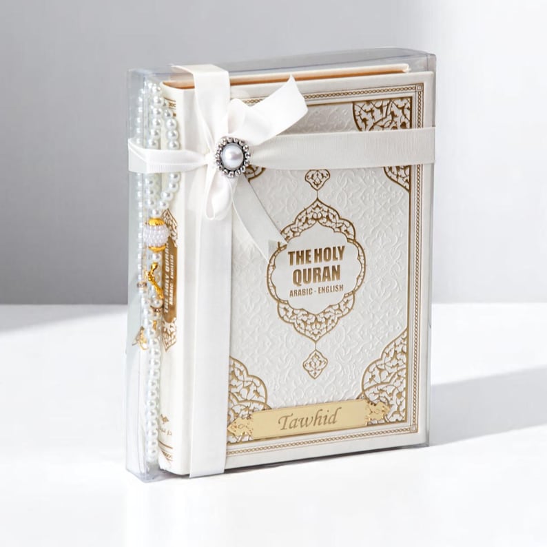 May include: A white and gold embossed book set, titled "The Holy Quran Arabic English," is presented in a clear box. A white ribbon with a pearl embellishment secures the set, which includes a prayer bead strand. The word "Tawhid" is inscribed.