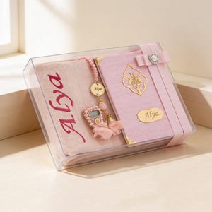 May include: A pink gift set in a clear box, featuring a prayer mat with "Alya" in red script, a pink book with gold accents, and a beaded rosary. The set includes a digital counter and is tied with a pink ribbon.