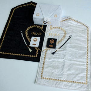Op de afbeelding: Een set huwelijksartikelen, waaronder een zwart en een wit gebedskleed met gouden randen en geborduurde namen. Een witte doos met de letters "O" en "B" en een klein boekje zijn ook inbegrepen. De items zijn op een wit oppervlak geplaatst.