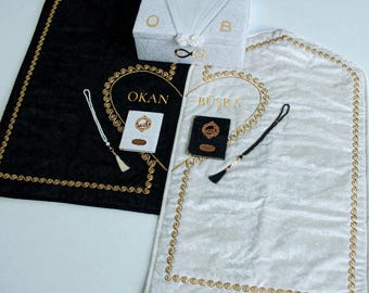 May include: A set of wedding items, including a black and a white prayer rug with gold trim and embroidered names. A white box with the letters "O" and "B" and a small book are also included. The items are arranged on a white surface.