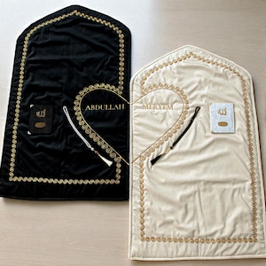 May include: Two prayer rugs, one black and one cream, are displayed. Both have gold embroidered borders and a heart-shaped design. Each rug includes a small book and a beaded tassel. The black rug has the name "ABDULLAH" and the cream rug has the name "MERYEM".