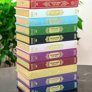 May include: A stack of colorful hardcover books titled "The Holy Quran" in Arabic and English. The books are in various colors, including red, blue, green, and purple, with gold lettering and decorative accents. The books are stacked on a dark surface.