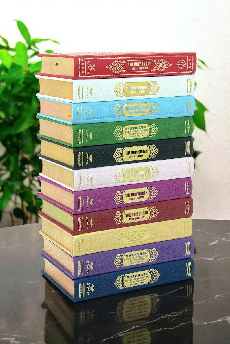 May include: A stack of colorful hardcover books titled "The Holy Quran" in Arabic and English. The books are in various colors, including red, blue, green, and purple, with gold lettering and decorative accents. The books are stacked on a dark surface.