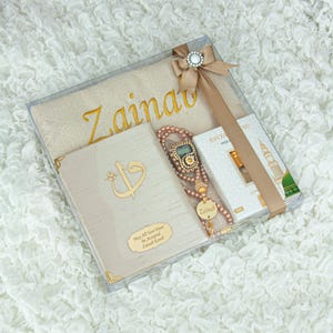 May include: A beige gift set in a clear box, featuring a prayer mat with gold lettering, a book with gold accents, a beaded tasbih, and a small perfume box. The set is tied with a beige ribbon and a decorative clasp.