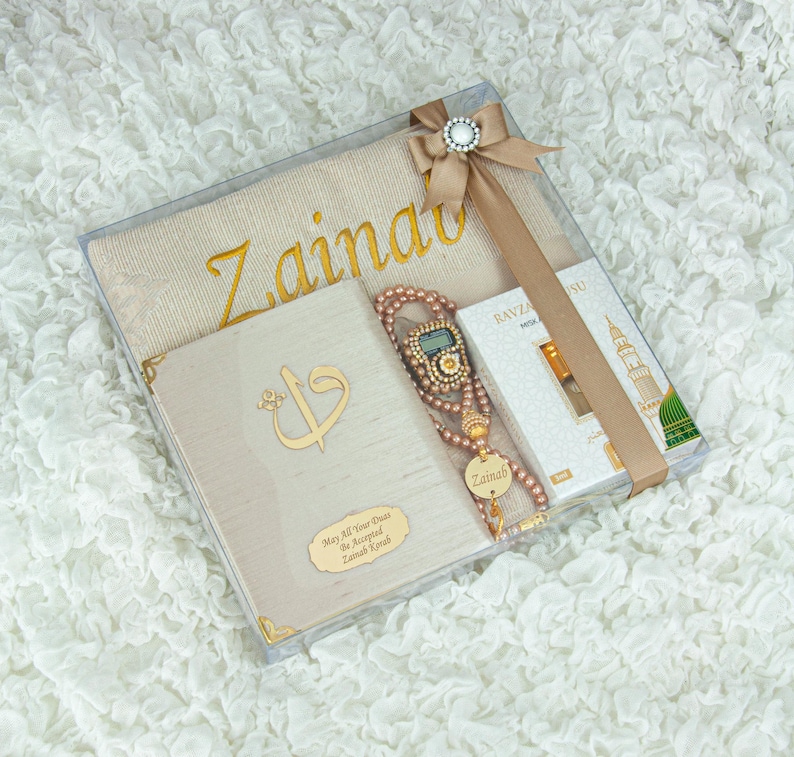 May include: A beige gift set in a clear box, featuring a prayer mat with gold lettering, a book with gold accents, a beaded tasbih, and a small perfume box. The set is tied with a beige ribbon and a decorative clasp.