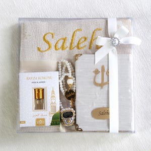 May include: A gift set in a clear box, featuring a beige prayer rug embroidered with "Saleh" in gold, a pearl necklace, a small perfume bottle, a digital counter, and a white book with gold accents. The set is tied with a white ribbon.