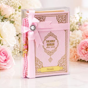 May include: A light pink Holy Quran book with gold foil lettering and ornate borders, encased in a clear plastic box. A pink satin ribbon with a pearl-centred bow wraps around the book, accompanied by a strand of pink prayer beads.