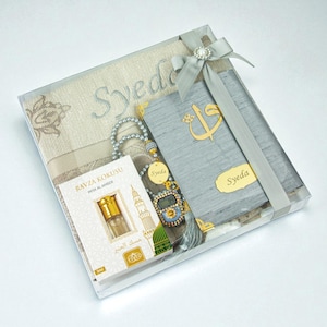 May include: A gift set in a clear box, including a prayer rug, a book, a digital prayer bead counter, and a small bottle of perfume. The set is tied with a grey ribbon and features the name "Syeda" on several items.