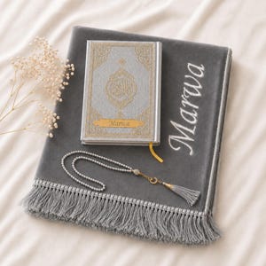 Op de afbeelding: Een grijze gebedskleed met franjes, een zilverkleurige Koran met gouden accenten en de naam "Marwa" op een gouden label, en een kralenrozenkrans. De Koran heeft goudkleurige reliëfdesigns. De mat heeft de naam "Marwa" erop geborduurd.
