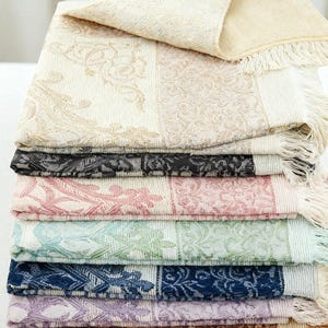 May include: A stack of patterned throws in cream, black, pink, blue, and purple. Each throw has a floral design and fringed edges. The top throw is folded over, showing its underside. The throws are suitable for a sofa or bed.