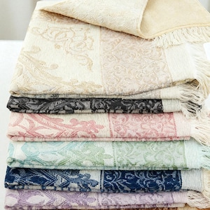 May include: A stack of decorative, patterned throws in various colors, including cream, black, pink, blue, and purple. Each throw features a floral design and fringe along the edges. The top throw is folded over, revealing a soft, textured surface.