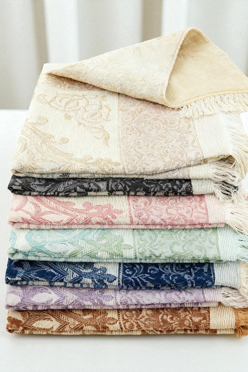 May include: A stack of decorative, patterned throws in various colors, including cream, black, pink, blue, and purple. Each throw features a floral design and fringe along the edges. The top throw is folded over, revealing a soft, textured surface.
