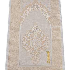 May include: Beige prayer rug with intricate gold-colored floral and geometric patterns. The rug features a central arched design and a decorative border. The word "Zainab" is written in gold script on the bottom right corner. The rug has a fringed edge.